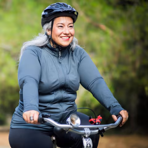 An adult smiling while cycling outdoors on a tree-lined trail.